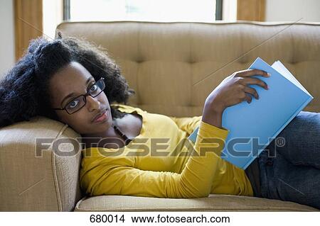 A young woman lying on a couch with a book, looking at camera View Large Photo Image Picture - A young woman lying on a couch with a book, looking at camera. Fotosearch