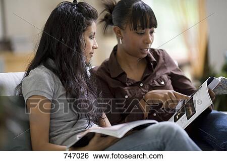 Stock Photograph - Two young woman looking at magazines on a sofa. Fotosearch