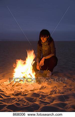 Stock Photograph - A woman staring into a fire burning on a beach. Fotosearch