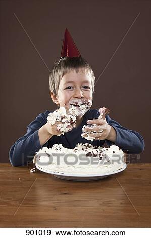 Stock Photo - A boy wearing a party hat eating pie with his hands, studio shot. Fotosearch