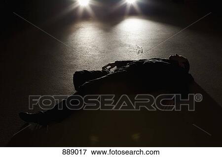 Stock Photo - A man lying on the ground in front of a car at night. Fotosearch