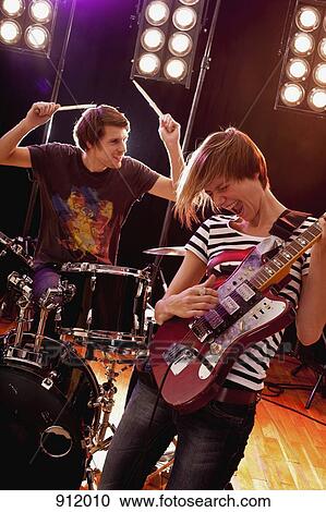 Stock Image - A man playing drums and a woman playing guitar in a rock band performing on stage. Fotosearch