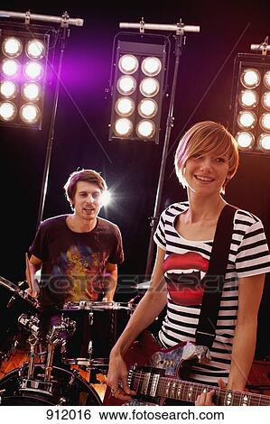 Stock Photograph - A man playing drums and a woman playing guitar in a rock band performing on stage. Fotosearch