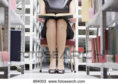 Stock Image - A woman sitting in a library with a book, focus on midsection and low section. Fotosearch