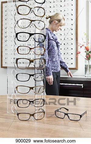 Stock Photo - An eyeglasses display in an eyewear store and a woman shopping in background. Fotosearch