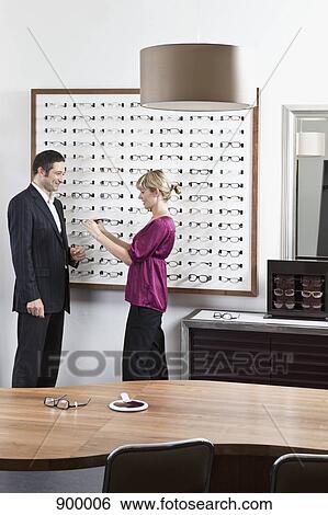 Stock Photograph - An optician holding up glasses to a man in an eyewear store. Fotosearch