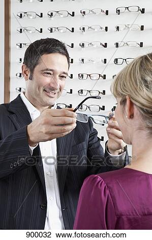 Stock Image - An optician holding up glasses to a customer in an eyewear store. Fotosearch