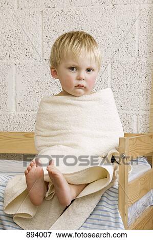 Stock Photo - An unhappy baby boy wrapped in a towel sitting on a bed. Fotosearch