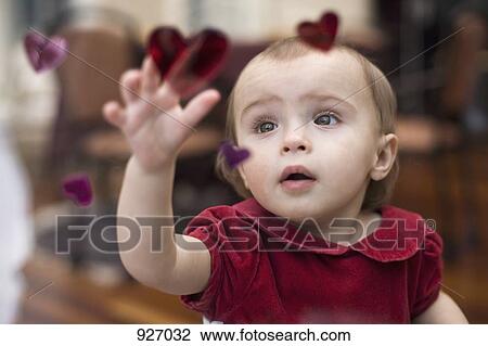 Stock Image - A baby reaching up to touch a heart decal window decoration, viewed through window. Fotosearch