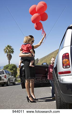 A woman holding a toddler and a bunch of balloons next to a girl getting into a car View Large Photo Image Stock Image - A woman holding a toddler and a bunch of balloons next to a girl getting into a car. Fotosearch
