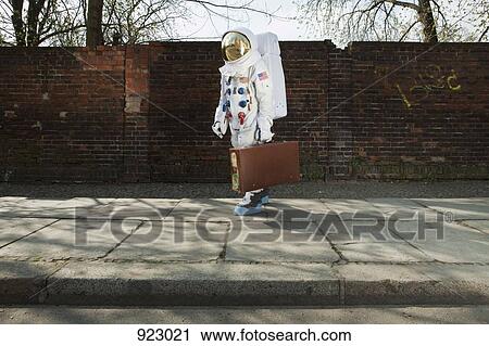 Stock Image - An astronaut carrying a suitcase and walking on a city sidewalk. Fotosearch