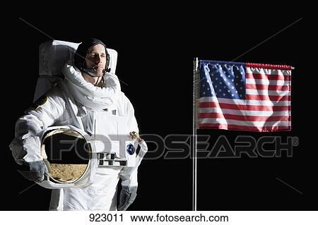 Stock Image - An astronaut holding his helmet standing next to an American flag, portrait. Fotosearch