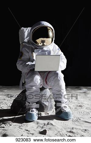 Stock Image - An astronaut on the moon sitting on a rock using a laptop. Fotosearch
