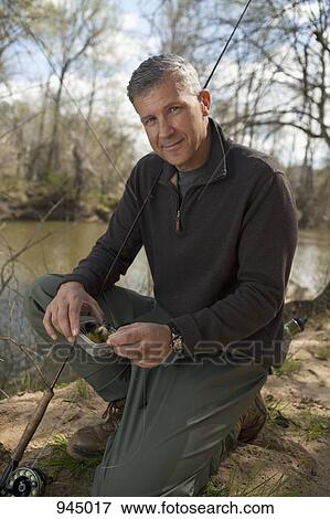 Stock Photo - Portrait of a man kneeling next to a river with fishing equipment. Fotosearch