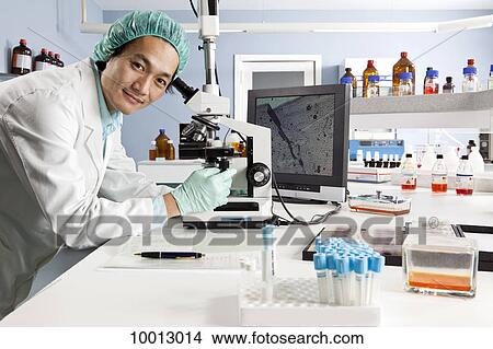 A lab technician using a microscope, looking at camera View Large Photo Image Picture - A lab technician using a microscope, looking at camera. Fotosearch