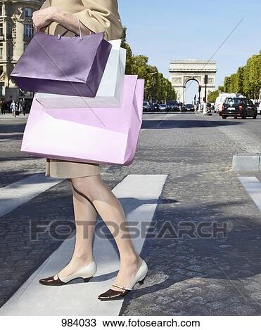 Stock Image - A woman carrying shopping bags walking across a zebra crossing, Paris, France. Fotosearch