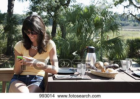 Picture - A woman using a smart phone while sitting at a table set for breakfast, outdoors. Fotosearch