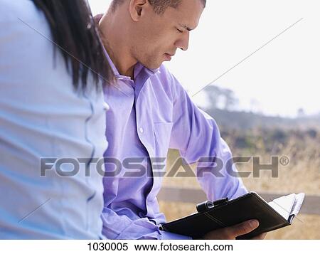 A businessman sitting next to a businesswoman reading his daily planner View Large Photo Image Stock Photography - A businessman sitting next to a businesswoman reading his daily planner. Fotosearch
