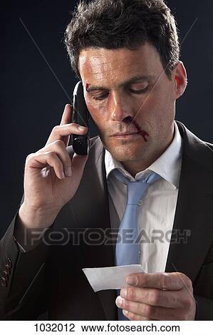Stock Image - A businessman with bruises and a bloody nose looking at a business card while on his mobile. Fotosearch