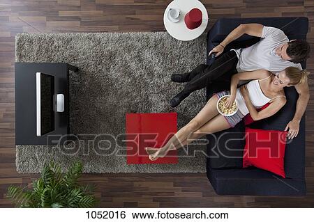 Stock Photograph - A cheerful couple watching TV and eating popcorn in their living room, overhead view. Fotosearch