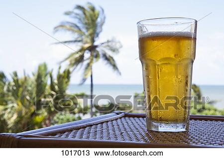 Stock Image - A glass of cold beer on a table with a view of a beach. Fotosearch