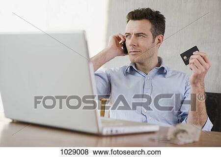 A man holding a credit card while on the phone and using a laptop View Large Photo Image Stock Photo - A man holding a credit card while on the phone and using a laptop. Fotosearch