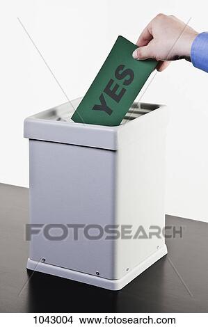 Picture - A man putting a ballot with the word YES written on it into a ballot box, close-up hands. Fotosearch