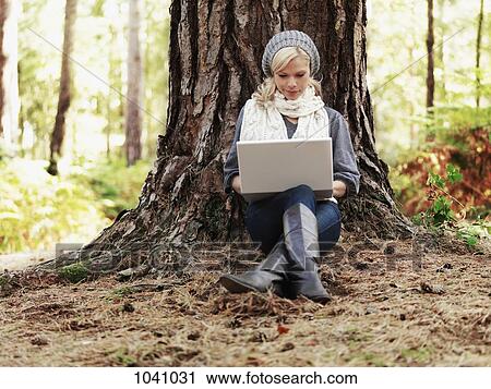 A woman in nature leaning against a tree and using a laptop View Large Photo Image Stock Image - A woman in nature leaning against a tree and using a laptop. Fotosearch