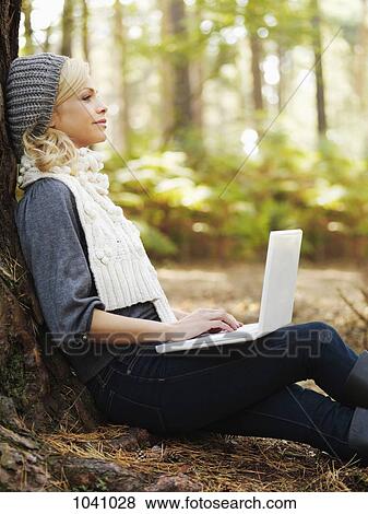 A woman in nature leaning against a tree and using a laptop View Large Photo Image Stock Photo - A woman in nature leaning against a tree and using a laptop. Fotosearch