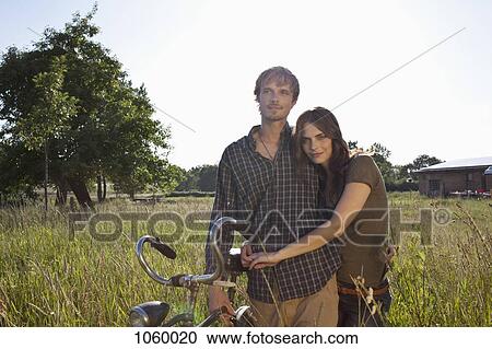 Couple with arms around each other walk through field with bike View Large Photo Image Stock Image - Couple with arms around each other walk through field with bike. Fotosearch