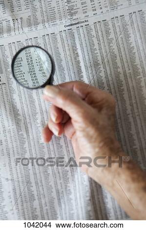 Picture - Detail of a senior woman holding a magnifying glass over newspaper stock pages. Fotosearch
