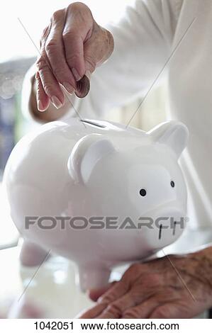 Stock Image - Detail of a senior woman putting a coin into a piggy bank. Fotosearch