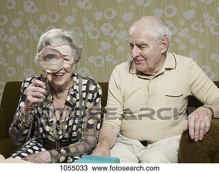 Stock Image - Senior man amused as senior woman magnifies appearance of her eye with magnifying glass. Fotosearch