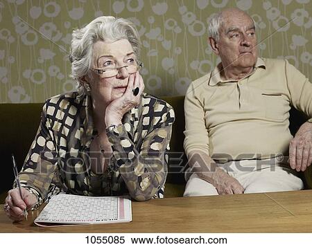 Senior woman works out magazine puzzle with senior man in background View Large Photo Image Stock Photography - Senior woman works out magazine puzzle with senior man in background. Fotosearch