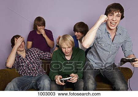 Stock Image - Two teenage boys playing a video game while their friends cheer them on. Fotosearch