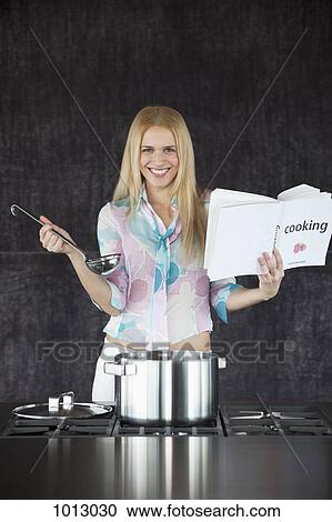 Stock Image - Woman cooking looking happy holding cookery book and ladle.. Fotosearch