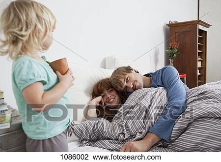 Stock Image - A couple lying in bed with their child standing beside the bed. Fotosearch