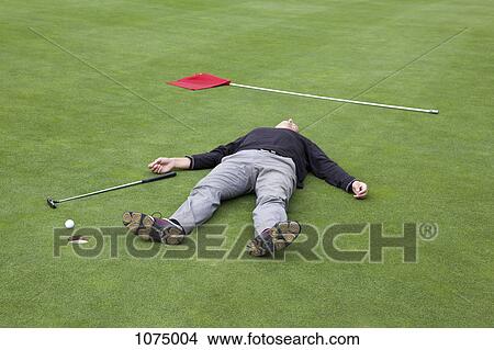 Picture - A distraught golfer lying on putting green with ball at the edge of hole. Fotosearch