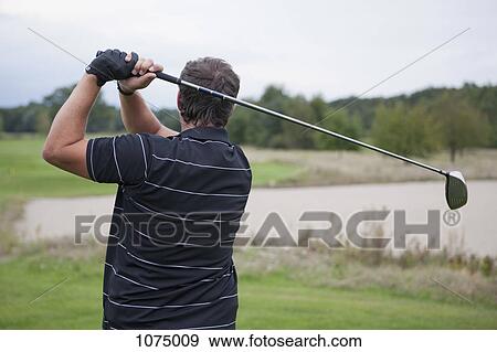Stock Photo - A golfer teeing off, rear view, waist up. Fotosearch