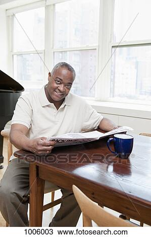 Stock Image - A mature man sitting at dining table holding a newspaper. Fotosearch
