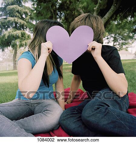 Stock Image - A teenage couple hiding behind a cut out heart. Fotosearch