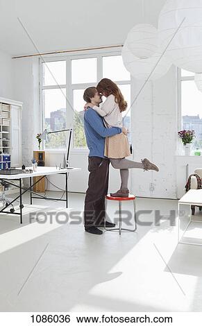 Stock Photograph - A woman standing on a stool with her arms around her boyfriend. Fotosearch