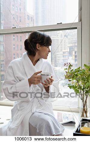 A woman wearing a bathrobe sitting on a window sill and holding a mug, looking at view View Large Photo Image Stock Image - A woman wearing a bathrobe sitting on a window sill and holding a mug, looking at view. Fotosearch