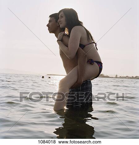 Stock Photo - A young man giving his girlfriend a piggyback ride in the sea. Fotosearch