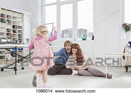 Picture - Parents sitting on floor watching their young daughter run towards them. Fotosearch