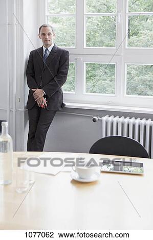 Stock Image - A businessman leaning against a window sill in a conference room. Fotosearch