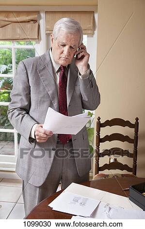 A businessman reading some paperwork and using a cell phone at home View Large Photo Image Stock Image - A businessman reading some paperwork and using a cell phone at home. Fotosearch