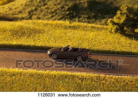 Stock Image - A diorama of a miniature man driving a toy convertible sports car on a rural road. Fotosearch