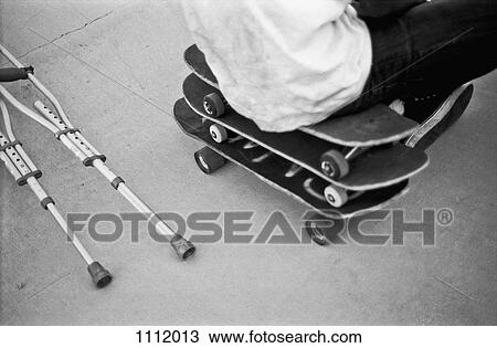 Stock Image - A man sitting on a stack of skateboards next to crutches. Fotosearch