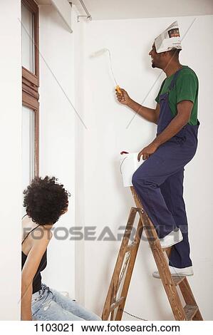 Stock Image - A man standing on a ladder using a paint roller while a woman watches. Fotosearch